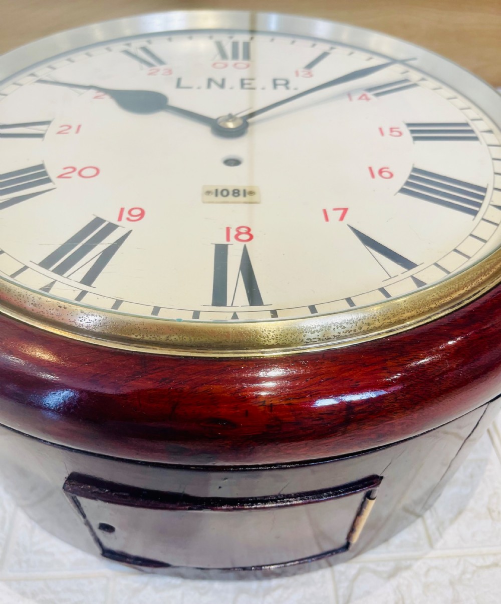 A circular LNER fusee railway wall clock featuring a dark mahogany wooden surround, brass bezel, cream dial with black Roman numerals and red 24-hour numbers. View XI