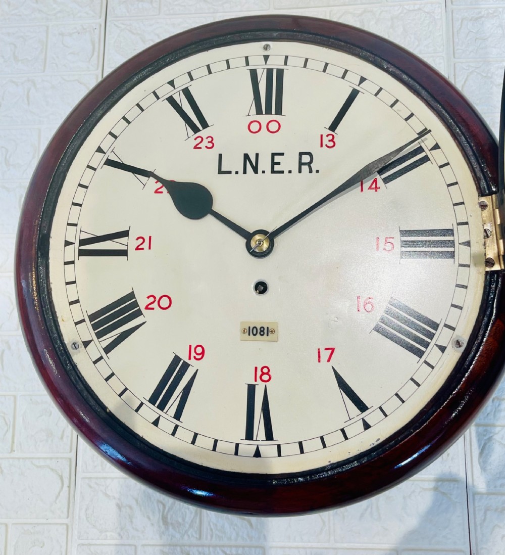 A circular LNER fusee railway wall clock featuring a dark mahogany wooden surround, brass bezel, cream dial with black Roman numerals and red 24-hour numbers. View XII