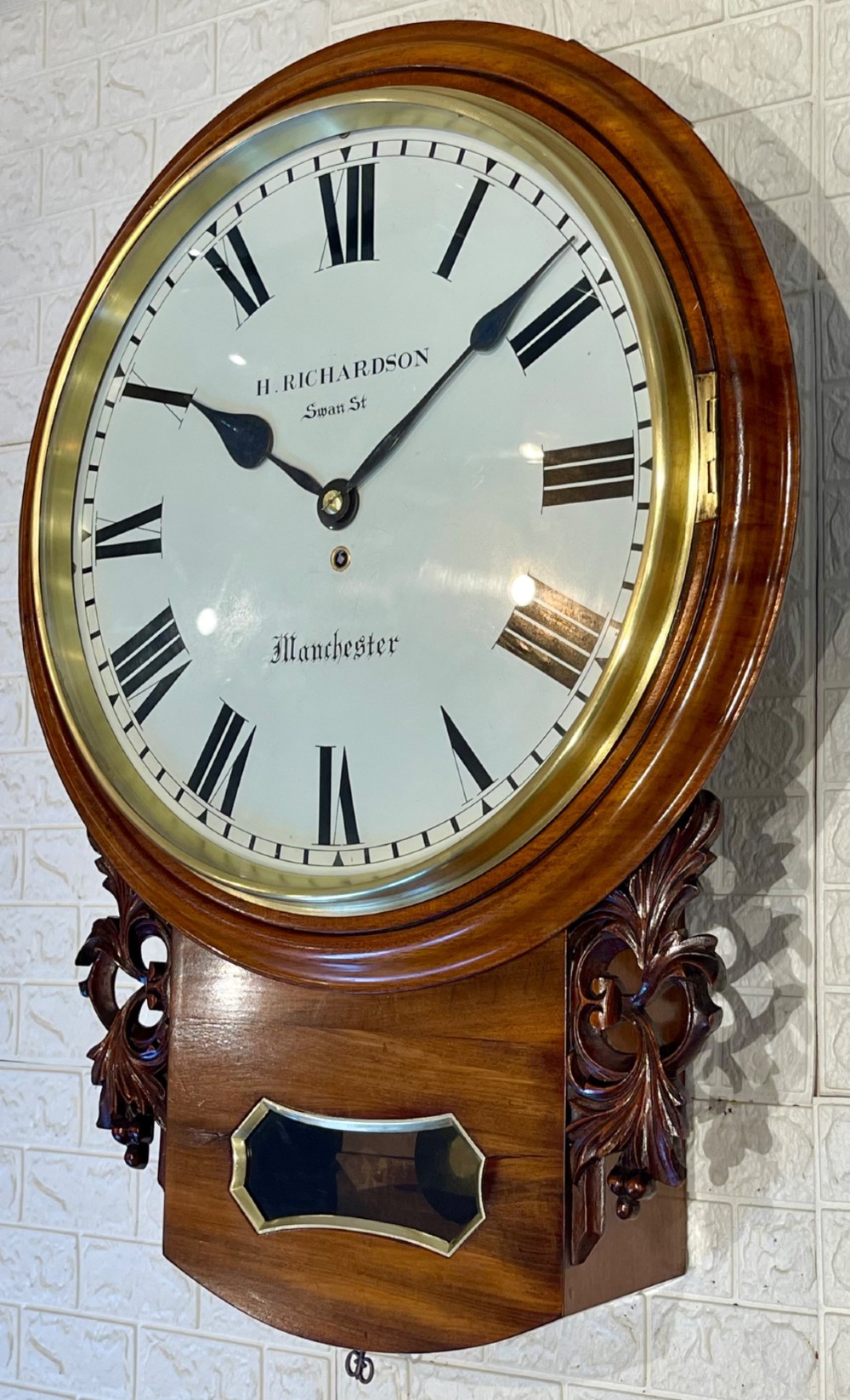Victorian oak drop dial wall clock featuring a circular white dial with Roman numerals, a brass bezel, and carved side scrolls. View II