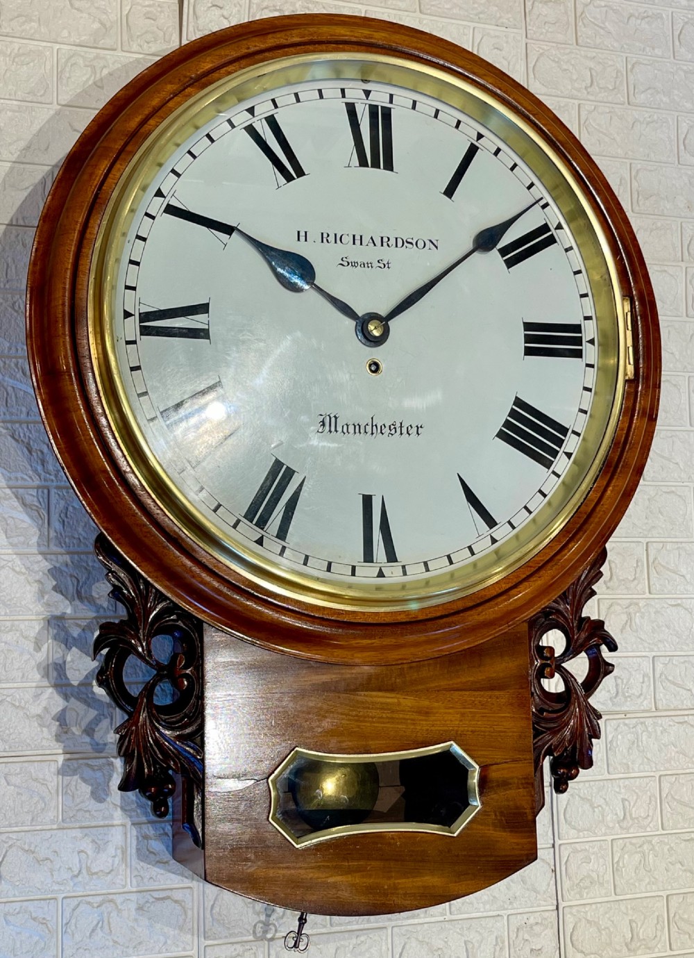 Victorian oak drop dial wall clock featuring a circular white dial with Roman numerals, a brass bezel, and carved side scrolls. View IV