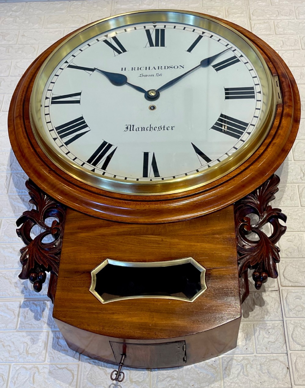 Victorian oak drop dial wall clock featuring a circular white dial with Roman numerals, a brass bezel, and carved side scrolls. View X