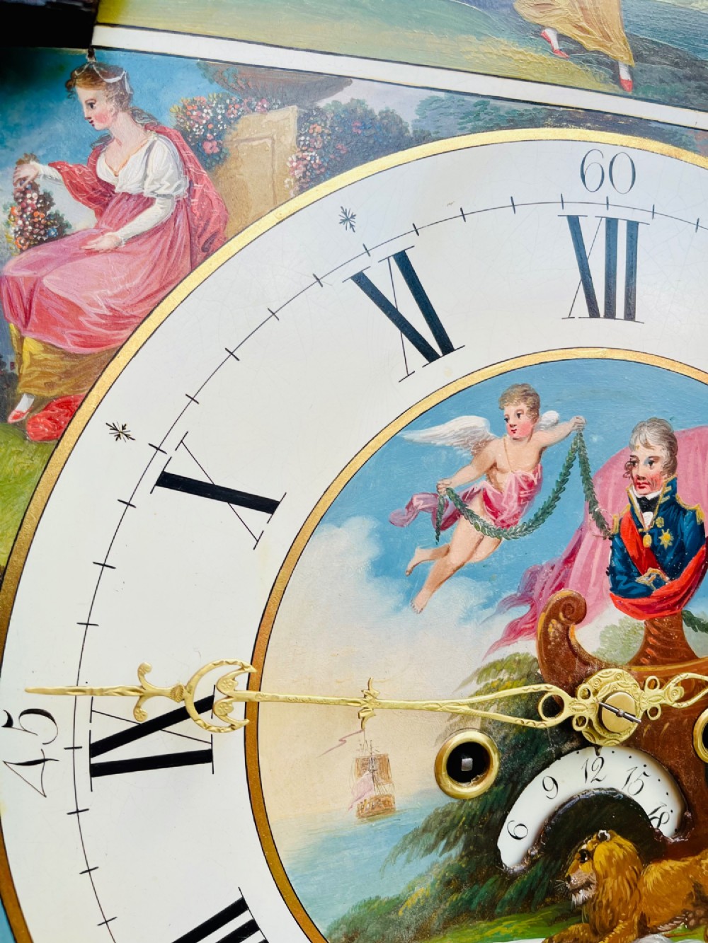 A tall Regency mahogany longcase clock featuring a swan-neck hood pediment, brass finial, fluted columns, and a hand-painted dial with figurative scenes. View X