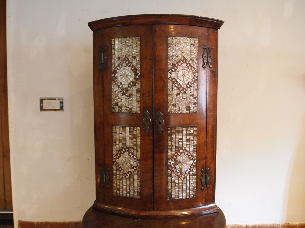 A dark walnut bowfront corner cabinet featuring two doors detailed with geometric mother of pearl inlay and brass hinges.