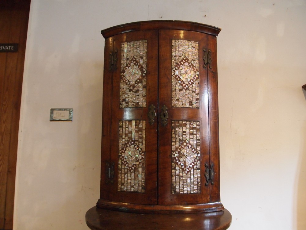 A dark walnut bowfront corner cabinet featuring two doors detailed with geometric mother of pearl inlay and brass hinges. View VI