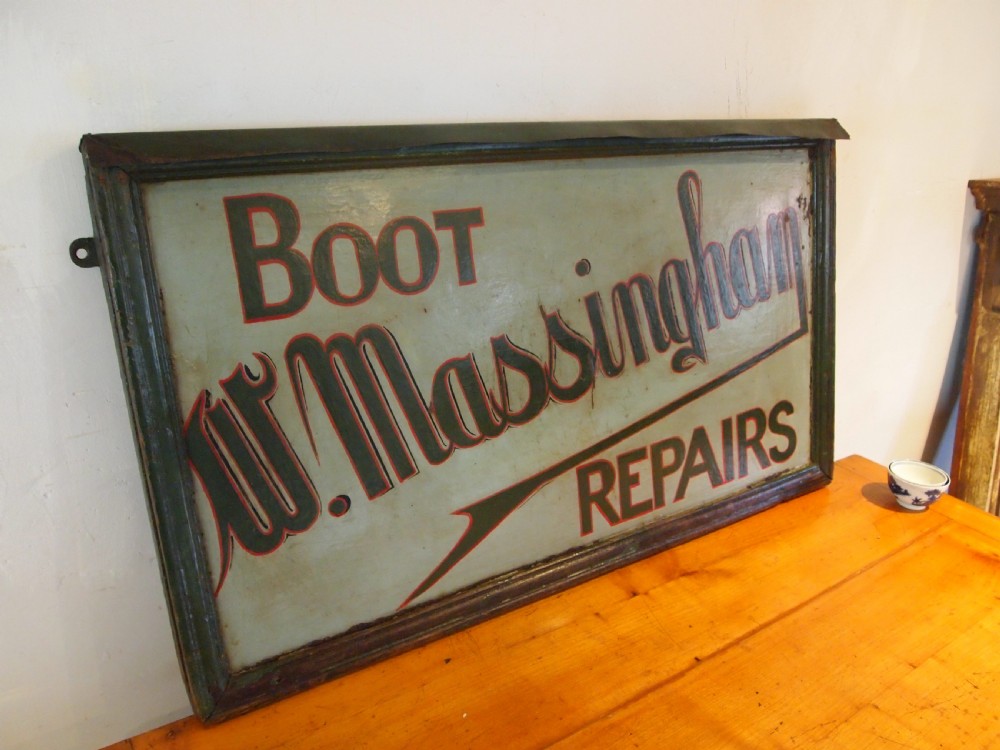 A rectangular hand-painted antique metal shop sign featuring black lettering outlined in red reading Boot W Massingham Repairs against a grey background within a dark metal frame.