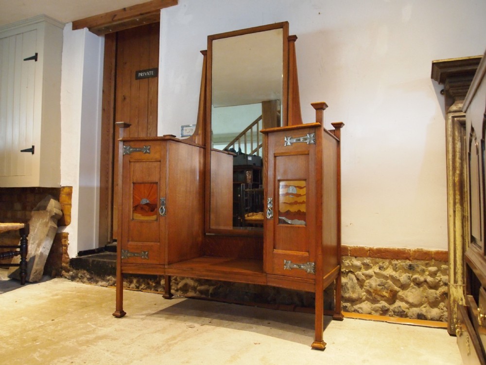 An Arts and Crafts solid oak dressing table featuring a central tall mirror flanked by two side cupboards with decorative inlaid landscape panels and metal strap hinges.