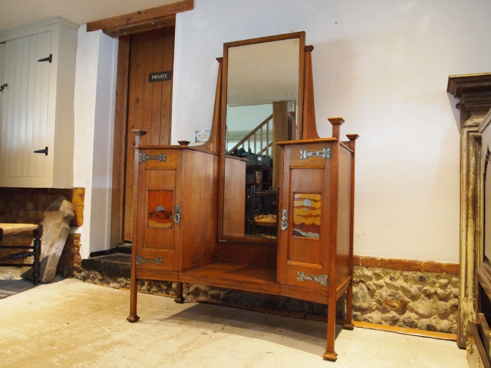 An Arts and Crafts solid oak dressing table featuring a central tall mirror flanked by two side cupboards with decorative inlaid landscape panels and metal strap hinges. View II
