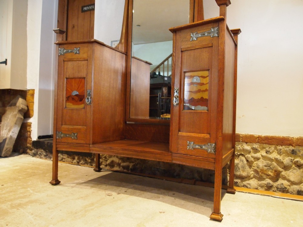An Arts and Crafts solid oak dressing table featuring a central tall mirror flanked by two side cupboards with decorative inlaid landscape panels and metal strap hinges. View VI
