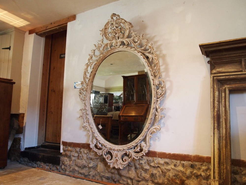 An 18th-century oval wall mirror featuring a heavily carved off-white wooden frame with acanthus leaf motifs and an ornate scrollwork cresting.