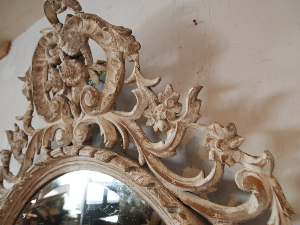An 18th-century oval wall mirror featuring a heavily carved off-white wooden frame with acanthus leaf motifs and an ornate scrollwork cresting. View II