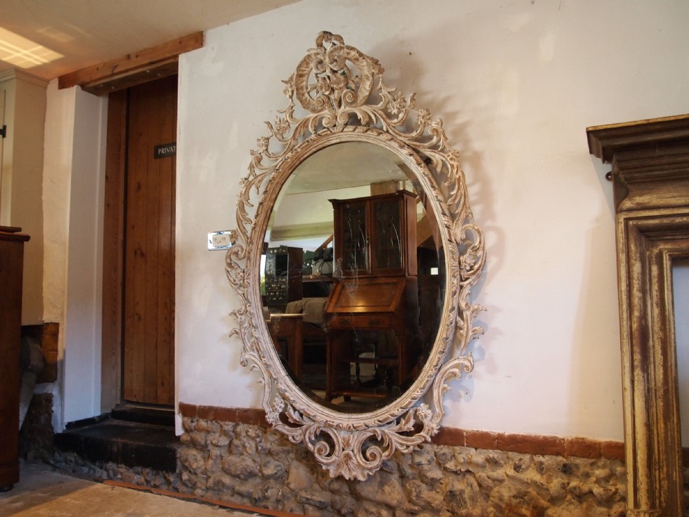 An 18th-century oval wall mirror featuring a heavily carved off-white wooden frame with acanthus leaf motifs and an ornate scrollwork cresting. View III