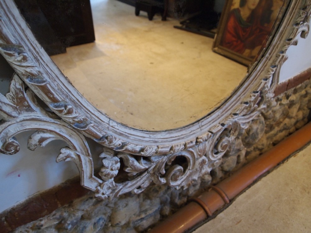 An 18th-century oval wall mirror featuring a heavily carved off-white wooden frame with acanthus leaf motifs and an ornate scrollwork cresting. View VI