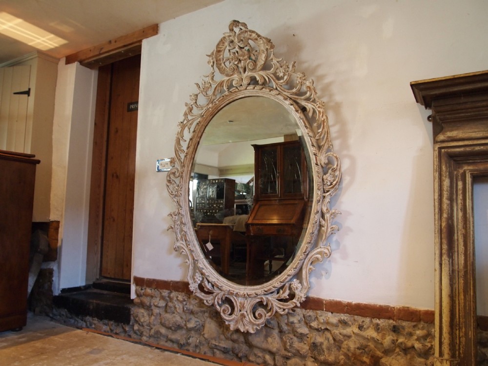 An 18th-century oval wall mirror featuring a heavily carved off-white wooden frame with acanthus leaf motifs and an ornate scrollwork cresting. View VIII