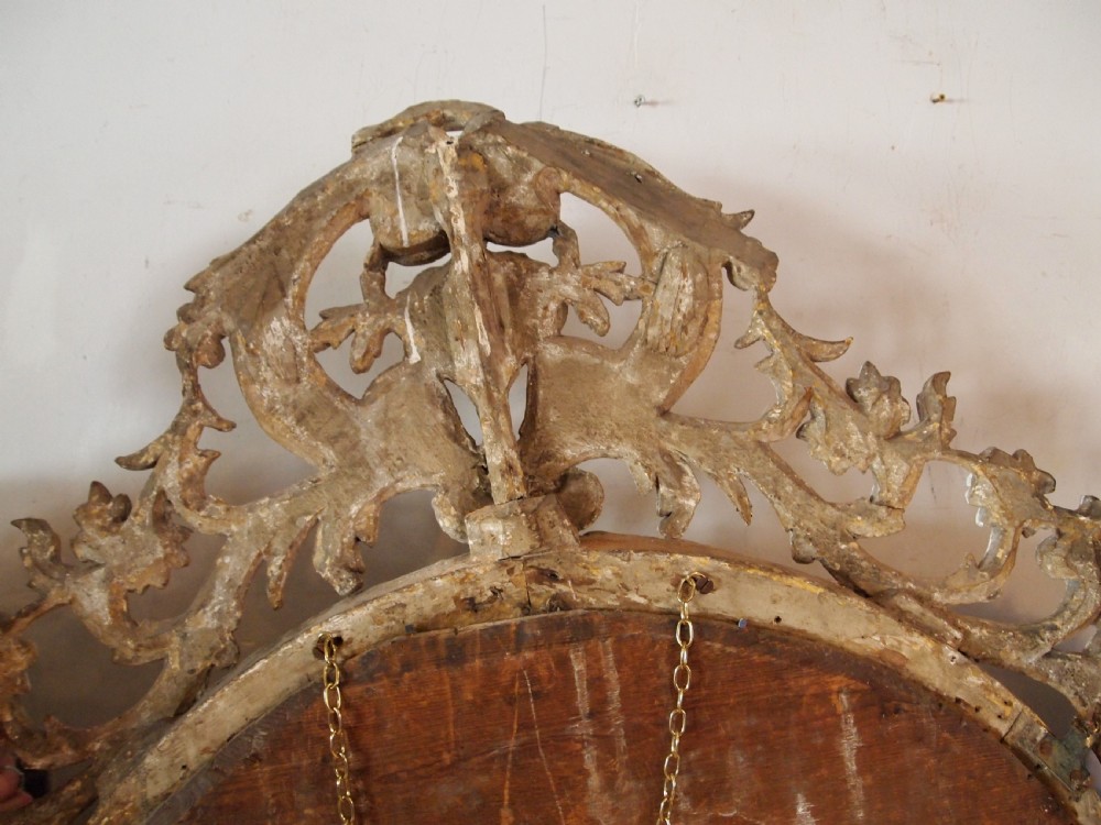 An 18th-century oval wall mirror featuring a heavily carved off-white wooden frame with acanthus leaf motifs and an ornate scrollwork cresting. View IX
