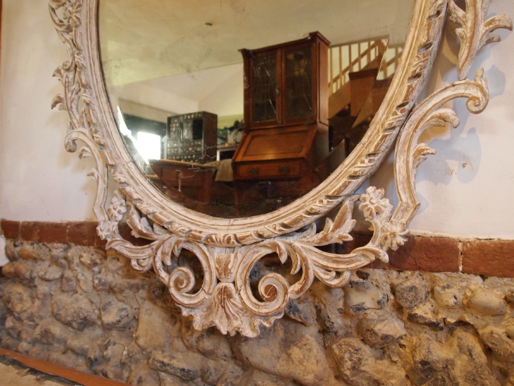 An 18th-century oval wall mirror featuring a heavily carved off-white wooden frame with acanthus leaf motifs and an ornate scrollwork cresting. View XI