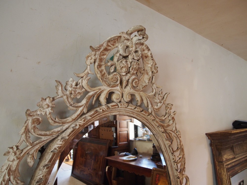 An 18th-century oval wall mirror featuring a heavily carved off-white wooden frame with acanthus leaf motifs and an ornate scrollwork cresting. View XII
