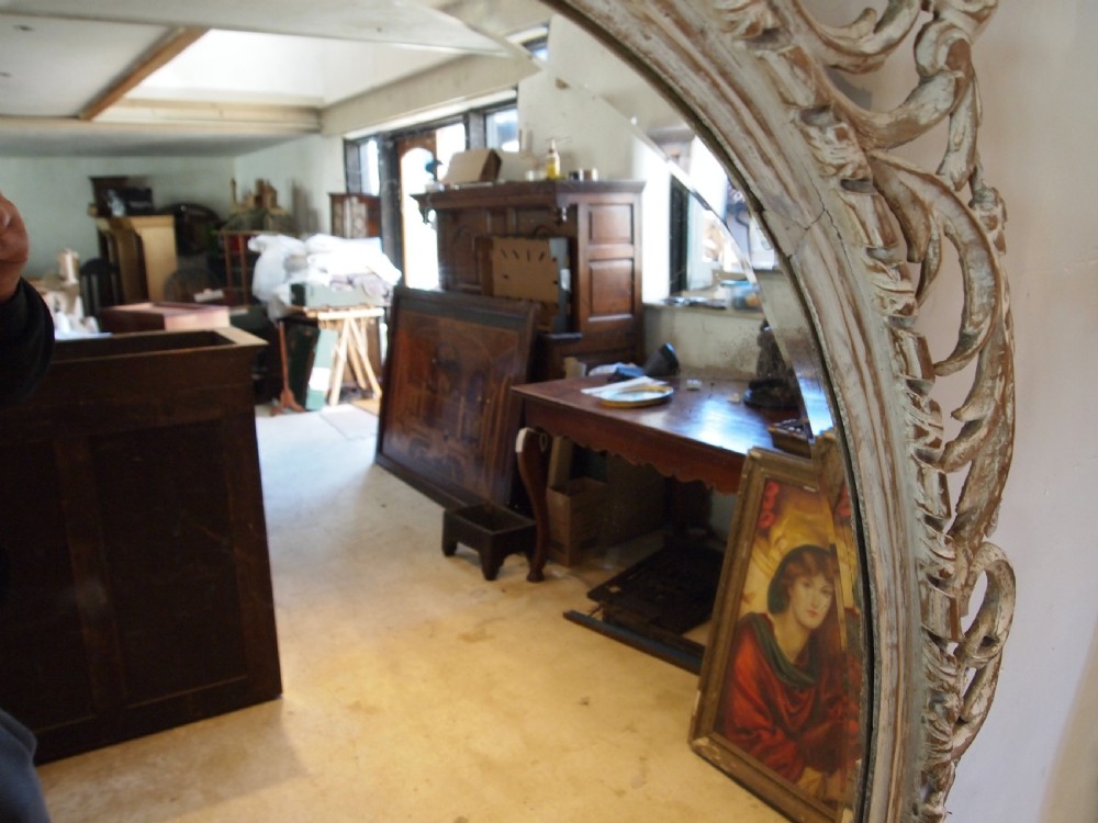 An 18th-century oval wall mirror featuring a heavily carved off-white wooden frame with acanthus leaf motifs and an ornate scrollwork cresting. View XIII