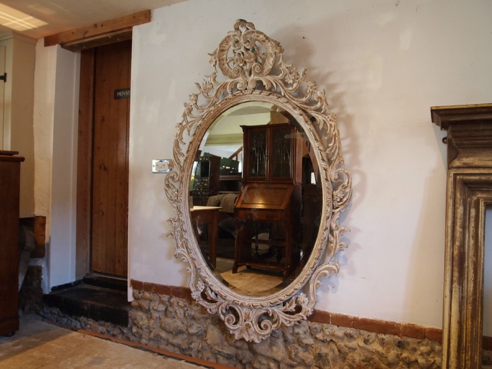 An 18th-century oval wall mirror featuring a heavily carved off-white wooden frame with acanthus leaf motifs and an ornate scrollwork cresting. View XV