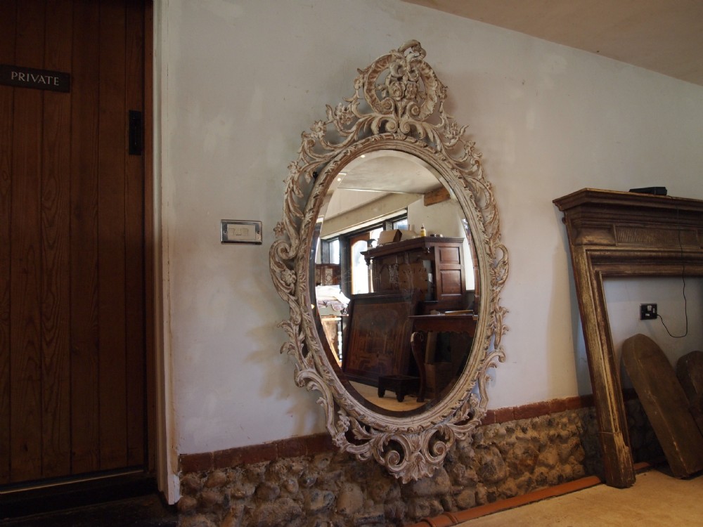 An 18th-century oval wall mirror featuring a heavily carved off-white wooden frame with acanthus leaf motifs and an ornate scrollwork cresting. View XVII