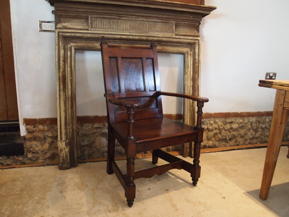 An eighteenth-century dark oak wainscot armchair featuring a panelled backrest, carved finials, turned front legs, and sturdy wooden stretchers.