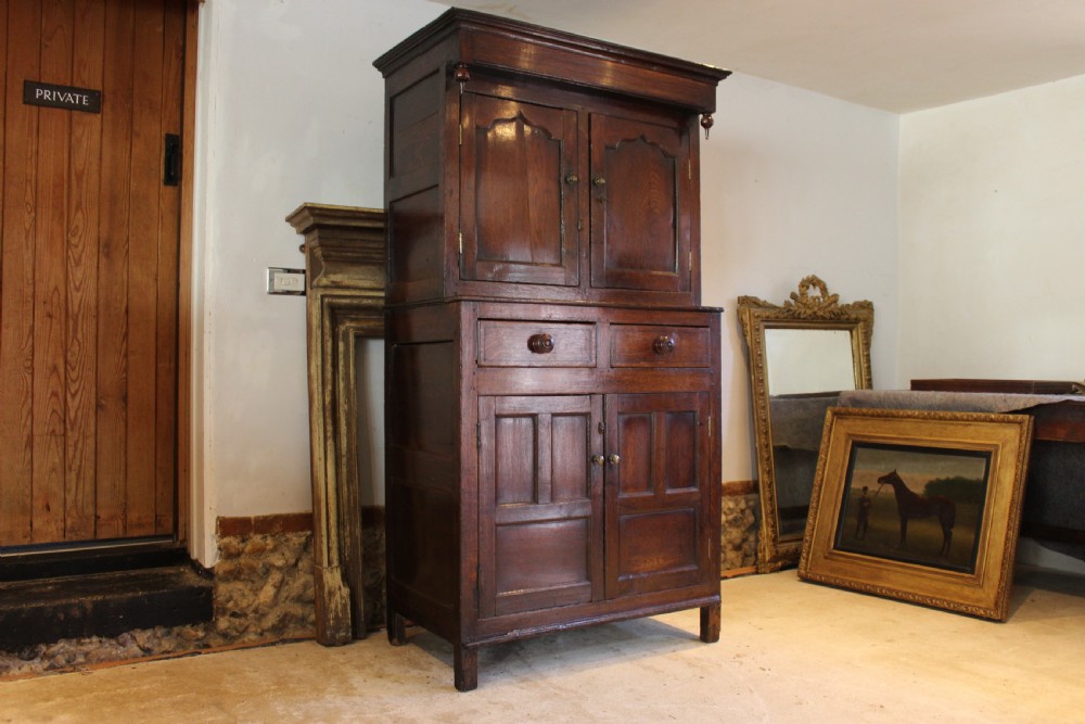 An 18th-century dark oak Welsh Cwpwrdd Deuddarn featuring a two-stage cupboard design with arched panelled upper doors, pendant drop finials, and lower drawers.
