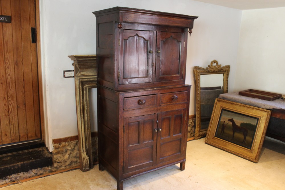An 18th-century dark oak Welsh Cwpwrdd Deuddarn featuring a two-stage cupboard design with arched panelled upper doors, pendant drop finials, and lower drawers. View X