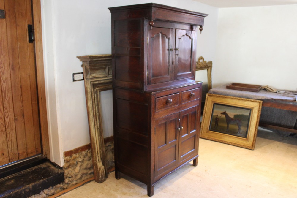 An 18th-century dark oak Welsh Cwpwrdd Deuddarn featuring a two-stage cupboard design with arched panelled upper doors, pendant drop finials, and lower drawers. View XIV