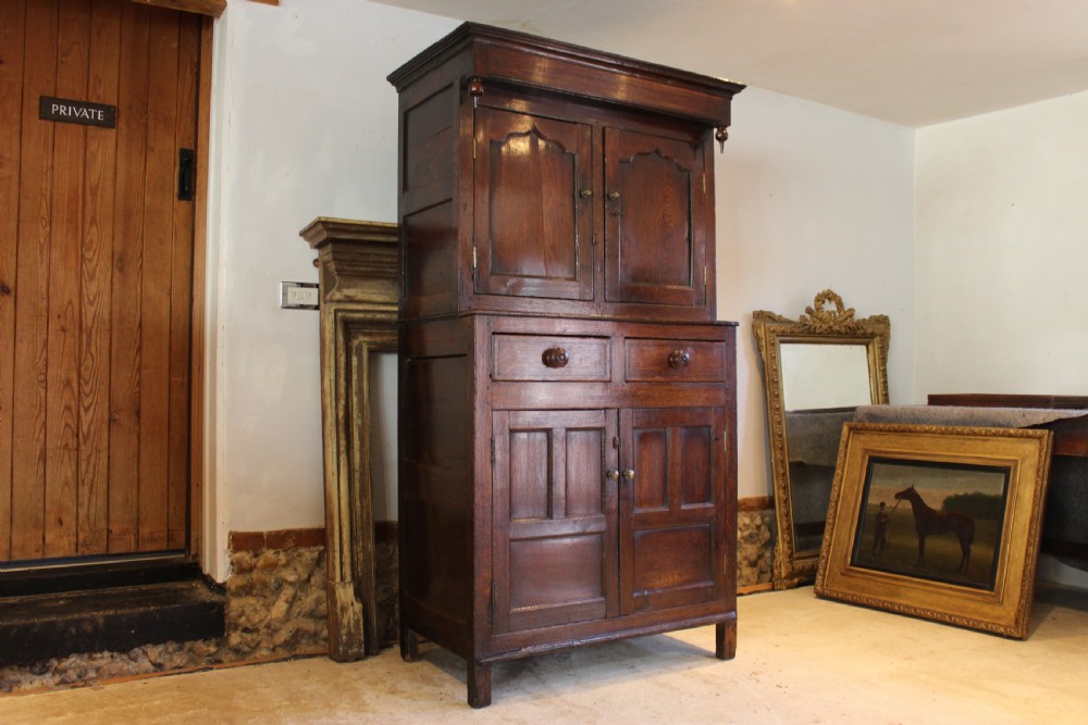 An 18th-century dark oak Welsh Cwpwrdd Deuddarn featuring a two-stage cupboard design with arched panelled upper doors, pendant drop finials, and lower drawers. View XVIII