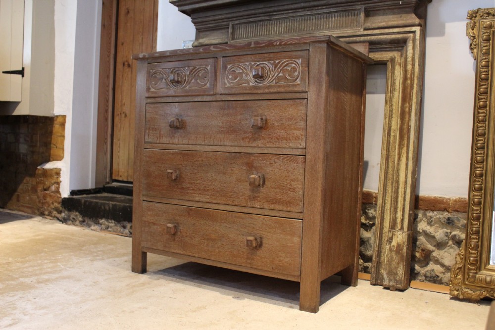 An Arts and Crafts limed oak chest of drawers featuring two upper drawers with stylised carved relief patterns and three long drawers below with square wooden handles.