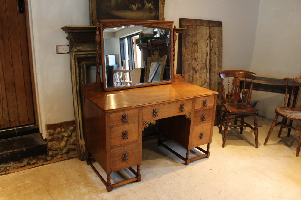 An Arts and Crafts solid oak dressing table featuring a central swinging mirror, carved spandrels, seven drawers with drop handles, and a kneehole stretcher base.