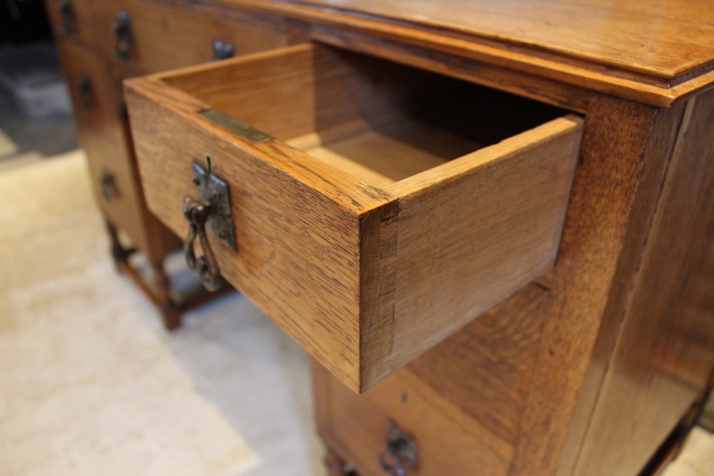 An Arts and Crafts solid oak dressing table featuring a central swinging mirror, carved spandrels, seven drawers with drop handles, and a kneehole stretcher base. View VII