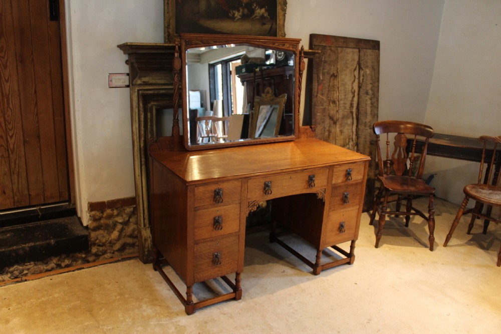 An Arts and Crafts solid oak dressing table featuring a central swinging mirror, carved spandrels, seven drawers with drop handles, and a kneehole stretcher base. View X
