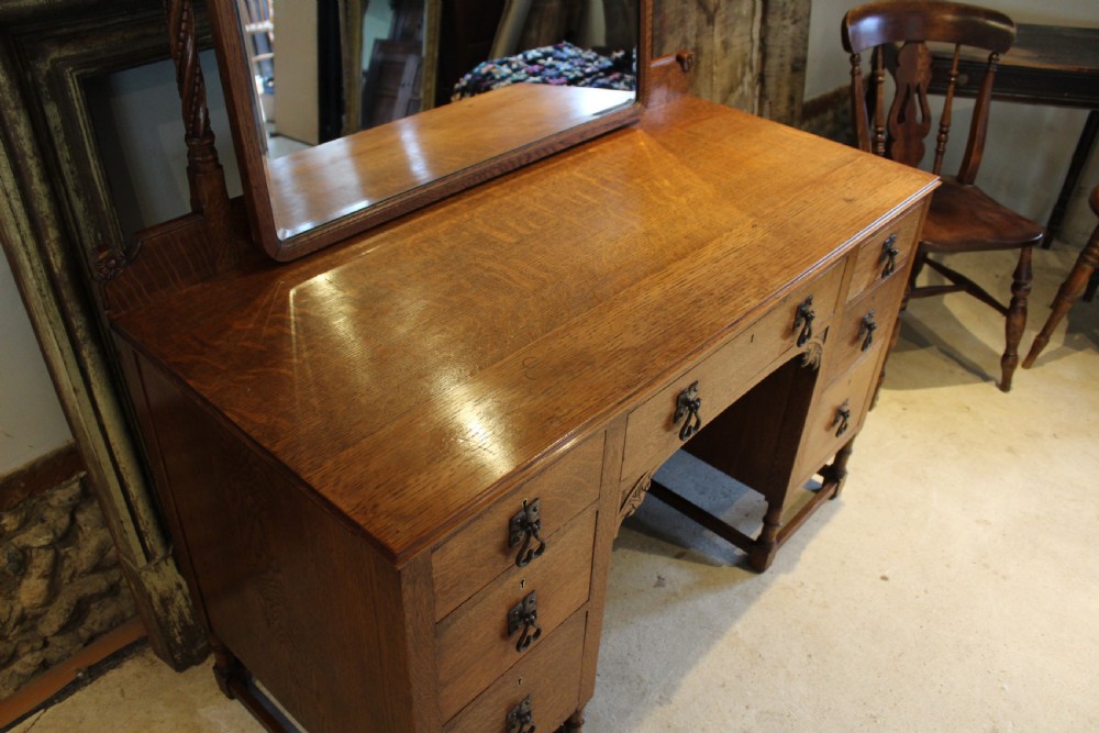 An Arts and Crafts solid oak dressing table featuring a central swinging mirror, carved spandrels, seven drawers with drop handles, and a kneehole stretcher base. View XI
