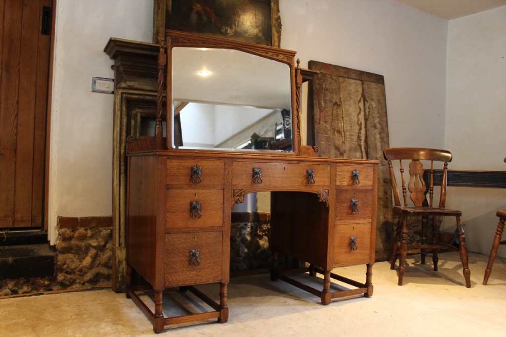 An Arts and Crafts solid oak dressing table featuring a central swinging mirror, carved spandrels, seven drawers with drop handles, and a kneehole stretcher base. View XII