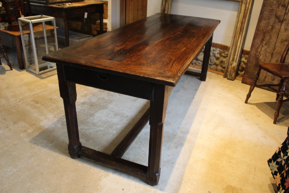 An 18th-century dark oak refectory dining table featuring a plank top, single end drawer, and square legs joined by a perimeter stretcher.