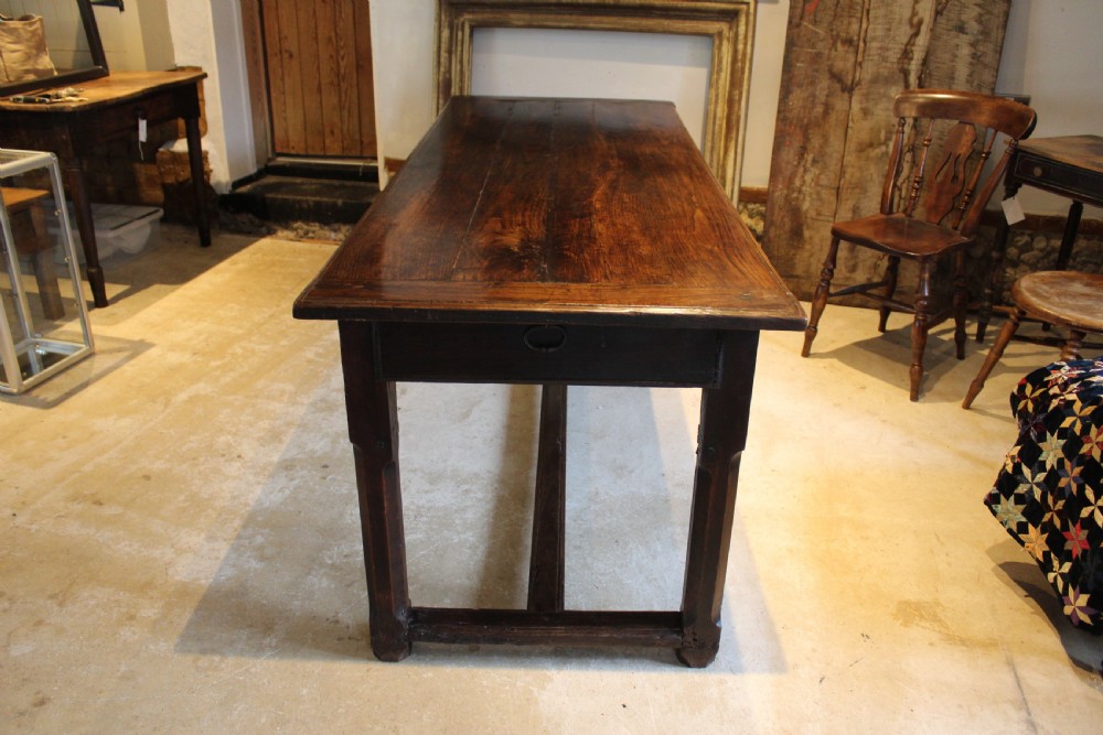 An 18th-century dark oak refectory dining table featuring a plank top, single end drawer, and square legs joined by a perimeter stretcher. View VIII