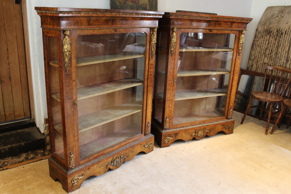 A pair of 19th-century Victorian walnut display cabinets featuring glazed single doors, adjustable interior shelves, and ornate gilt-bronze mounts on the corners and base.