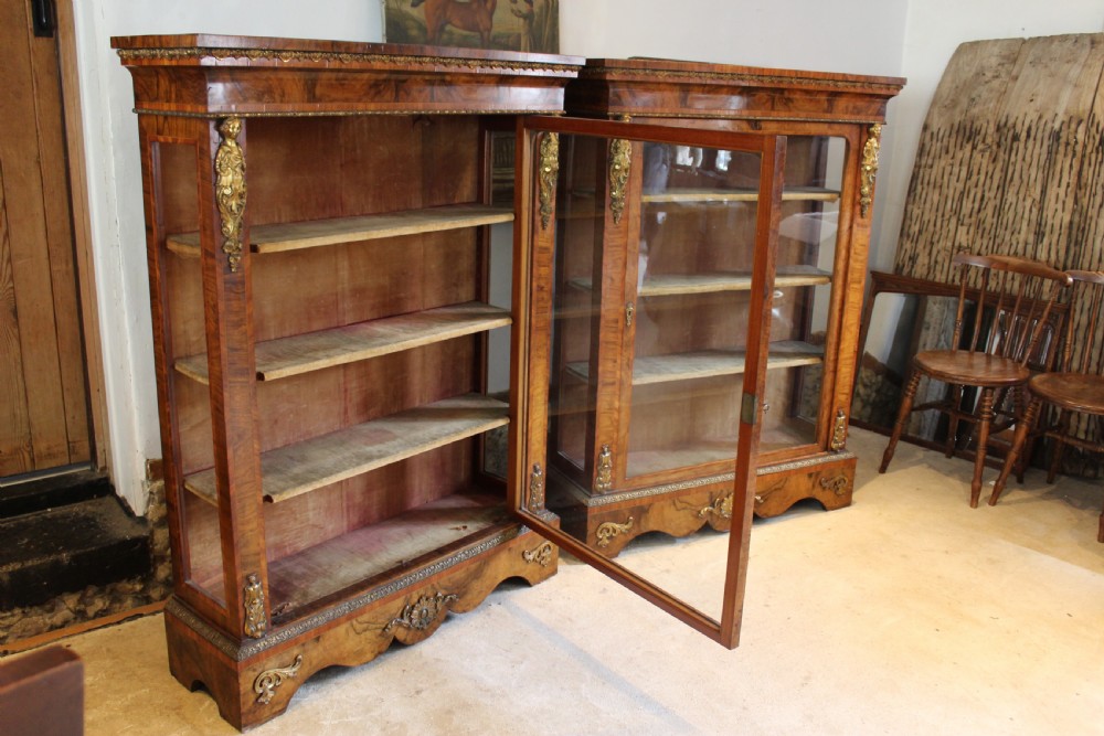 A pair of 19th-century Victorian walnut display cabinets featuring glazed single doors, adjustable interior shelves, and ornate gilt-bronze mounts on the corners and base. View VI