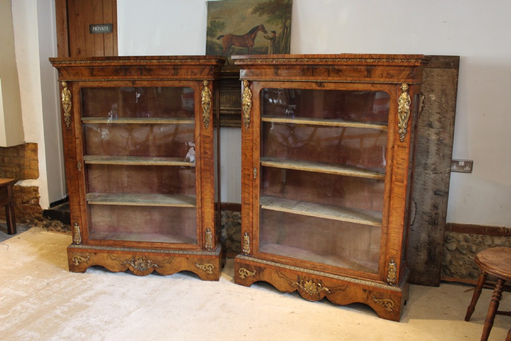 A pair of 19th-century Victorian walnut display cabinets featuring glazed single doors, adjustable interior shelves, and ornate gilt-bronze mounts on the corners and base. View X