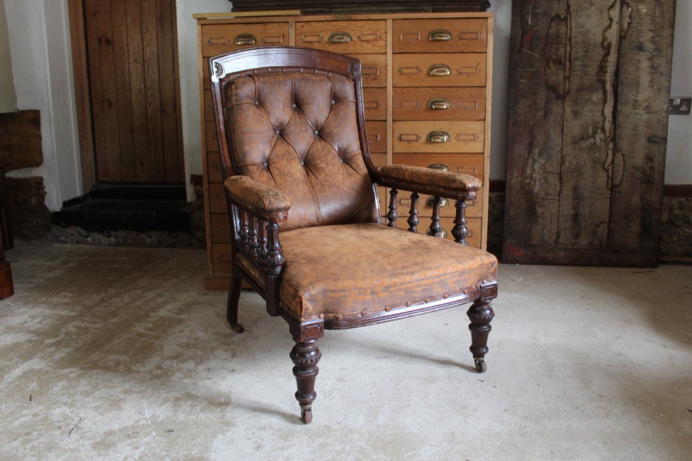 A late Victorian walnut armchair featuring button-backed brown leather upholstery, turned spindle arm supports, stud detailing, and carved front legs on castors.