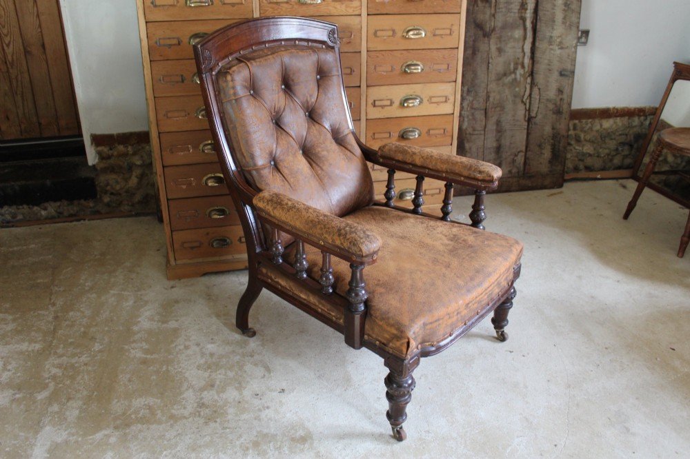 A late Victorian walnut armchair featuring button-backed brown leather upholstery, turned spindle arm supports, stud detailing, and carved front legs on castors. View II