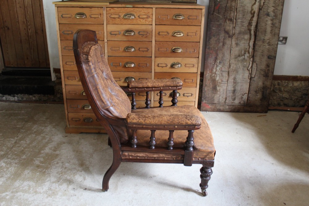 A late Victorian walnut armchair featuring button-backed brown leather upholstery, turned spindle arm supports, stud detailing, and carved front legs on castors. View IX