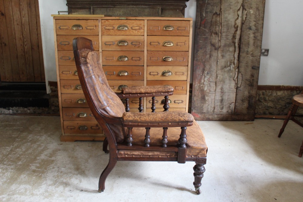 A late Victorian walnut armchair featuring button-backed brown leather upholstery, turned spindle arm supports, stud detailing, and carved front legs on castors. View X