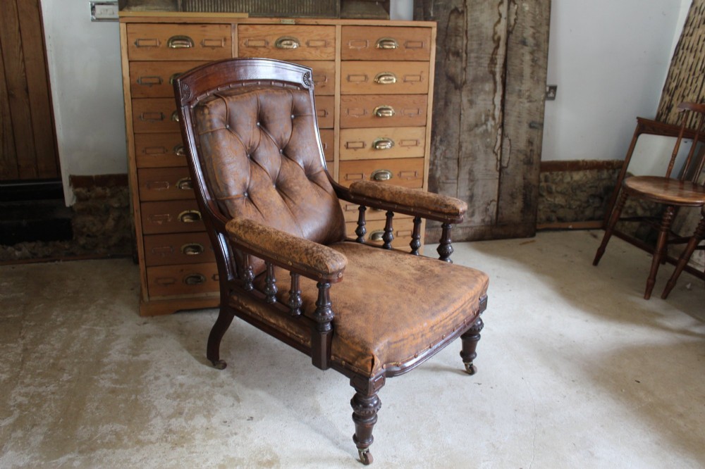 A late Victorian walnut armchair featuring button-backed brown leather upholstery, turned spindle arm supports, stud detailing, and carved front legs on castors. View XII