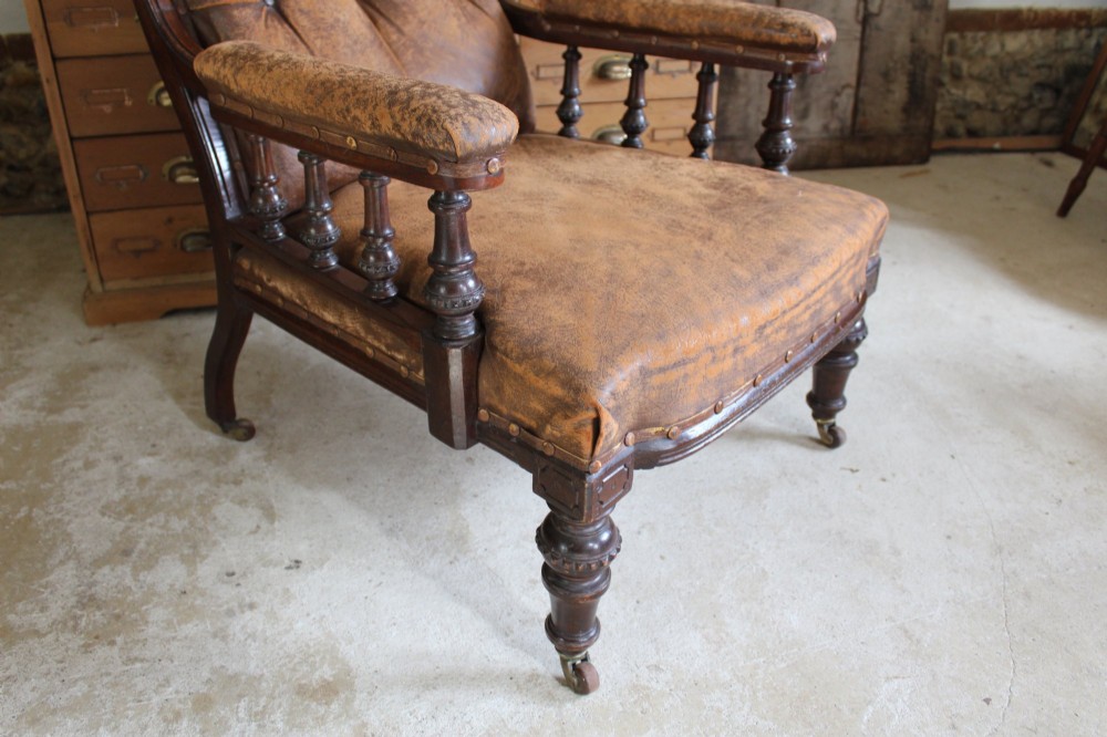 A late Victorian walnut armchair featuring button-backed brown leather upholstery, turned spindle arm supports, stud detailing, and carved front legs on castors. View XIII