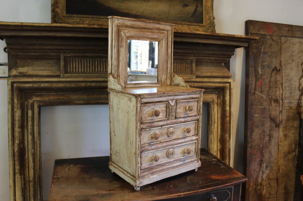 A late 19th-century French miniature wooden chest of drawers featuring a painted distressed cream finish, small turned wooden knobs, and an attached swivel mirror.