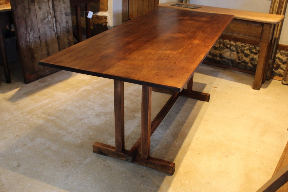 A solid teak Arts and Crafts refectory table featuring twin upright trestle supports resting on shoe feet and joined by a low stretcher rail.