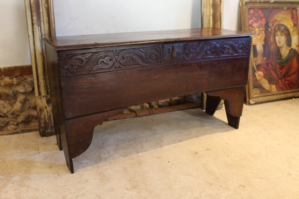 A dark elm six-plank coffer featuring a carved scrollwork frieze along the upper front rail, an iron escutcheon, and cutout boot-jack end supports.