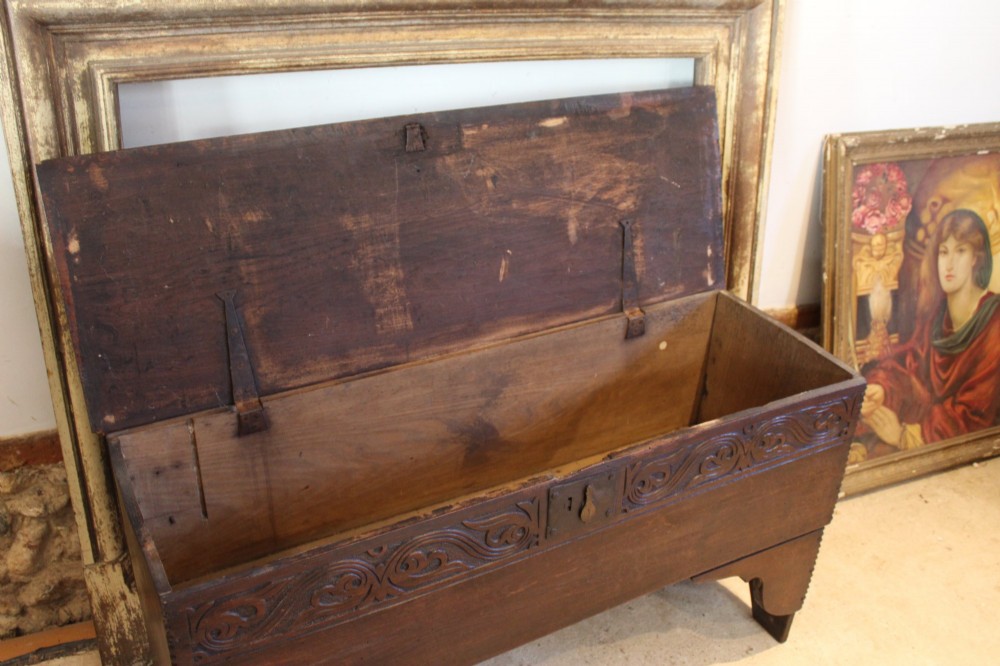 A dark elm six-plank coffer featuring a carved scrollwork frieze along the upper front rail, an iron escutcheon, and cutout boot-jack end supports. View VI