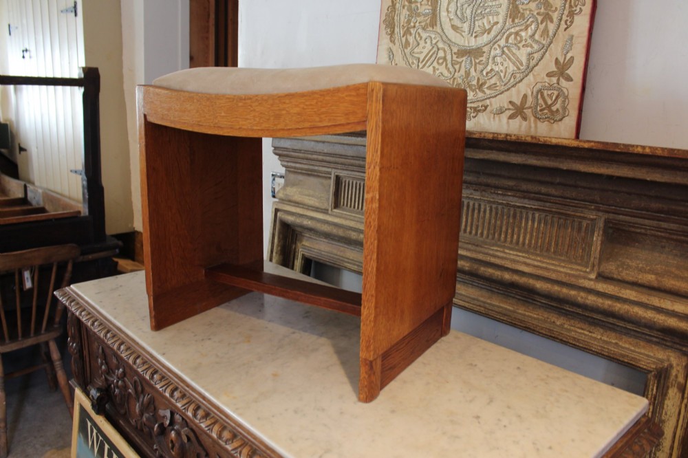 An oak stool featuring solid slab side supports, a central connecting stretcher, and a concave upholstered fabric seat cushion.